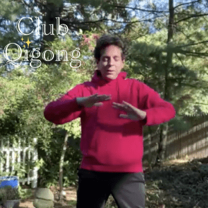 Joe practicing Qigong outdoors in a red hoodie, hands held gently in front of his stomach, surrounded by trees and greenery, with the words ‘Club Qigong ✨’ in the corner."